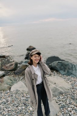 Rahat palto ve şapka giyen genç bir kadın bulutlu gökyüzünün altında sakin okyanus suyuyla kayalık sahilde huzurlu bir anın tadını çıkarıyor. Moda, doğa, sonbahar ve gülen modelli açık hava yaşam tarzı..