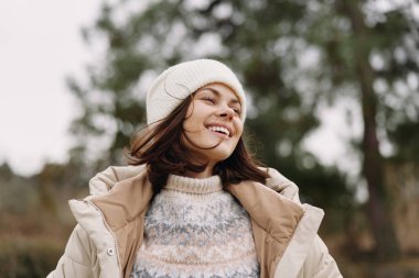 Açık havada, serin bir havada, örgü bir bere ve kalın bir ceket giymiş, havalı bir arka plan ve rahat bir yaşam tarzı ve mevsimsel moda havasında gülümseyen bir kadının portresi.