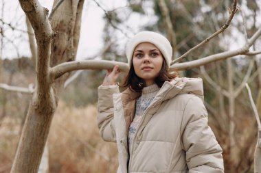 Açık hava sahnesinde yastıklı palto ve bereli bir kadın, yapraksız ağaçların arasında rahat bir poz, moda ve yaşam tarzı için kış havasında.