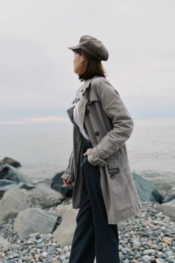 Kumaşlı palto ve şapka giyen genç bir kadın kayalık sahilde dikilip bulutlu havalarda sakin denizi seyrediyor. Sonbahar modası, açık hava yaşam tarzı, sakin ruh hali, doğa ve tefekkür kavramı..