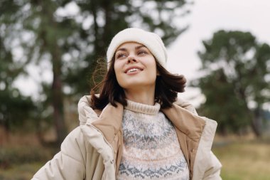 Açık havada ceketli ve bereli gülümseyen bir kadının portresi, arka planda ağaçlarla bir parkta dikilirken, tazeliği, özgürlüğü ve mevsimsel havayı anımsatıyor..