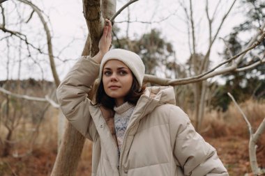 Bej rengi kışlık palto giymiş bir kadın portresi ve yapraksız ağaçların arasında bere, dingin ifade, serin tonlar, doğal ortam, mevsimsel hava ve sessiz yalnızlık, sakin bir açık hava görüntüsü.