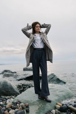 Koyu renk pantolon ve gri ceket giyen şık bir kadın bulutlu gökyüzü ile okyanus kenarındaki kayalık sahilde poz veriyor. Modern tarz kavramı, doğal manzara ve sıradan açık hava portresi..