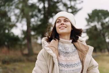 Açık hava sahnesinde bej ceketli, örgü kazaklı ve bereli gülümseyen bir kadın açık gökyüzü ve yapraklı arkaplanı olan bir parkta serin bir sonbahar gününün tadını çıkarıyor.