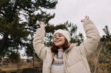 Sıcak bir palto giyen ve açık havada kazak ören mutlu bir kadın, özgürlüğünü kutluyor, ağaçların arasında kollarını açmış, açık havada..