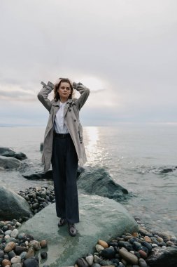 Uzun palto ve geniş pantolon giyen kendine güvenen kadın sakin denizin yanındaki kayalık sahilde duruyor. Bulutlu gökyüzü ve yumuşak doğal ışık sakin ve yansıtıcı atmosferi vurguluyor.