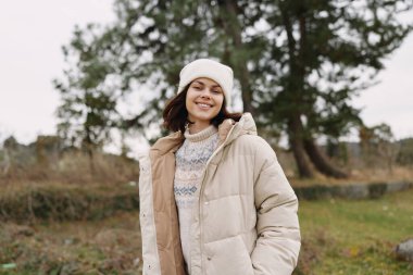 Kadın açık hava kışlık ceketi, ağaçlar ve çimenlerle dolu manzarada gülümseyen bereyle, sıcak ve rahat bir yaşam tarzı ve portre fotoğrafçılığı sergiliyor.