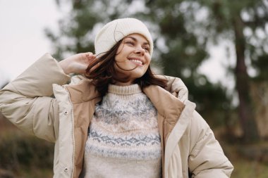 Sıcak bir palto içinde gülümseyen bir kadının portresi ve açık havada süveter örmek, serin havanın ve bulanık ağaçlarla kaplı bir parkta sıradan bir tarzın keyfini çıkarmak..