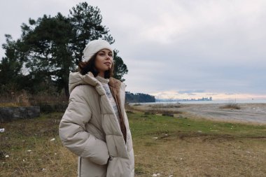 Sahilde yalnız bir kadının üzerinde rahat bir giysi, soğuk hava, engin sahil ve bulutlu gökyüzü, sakin doğal bir portre..