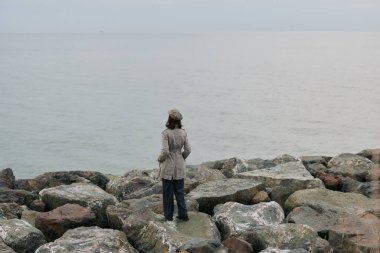 Palto ve şapka giyen kişi taş iskeleden sakin okyanusa bakıyor. Görüntü, bulutlu bir günde su kenarında yalnızlığı, yansımayı, doğayı ve sessiz atmosferi vurgular..