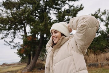 Açık havada, sıcak giyimli bir kadın, kollarını kaldırmış, soğuk havada gülümsüyor, ağaçlı ve çimenli doğa arkaplanı, rahat bir kış havası ve rahat bir sahne için..