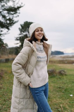 Kış modası, açık hava, rahat kıyafet, rahat ceket, günlük tarz, örgü ören kazaklı, bereli ve kotlu bir kadın bulutlu bir gökyüzünün altında açık bir arazide duruyor, rahat bir yaşam tarzı aktarıyor ve...