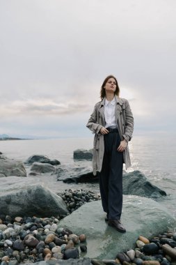 Palto ve pantolon giyen genç bir kadın su kenarında taş plajda poz veriyor. Kayalar ve çakıl taşlarıyla donuk bir arka plan. Açık hava moda imajı rahat ve düşünceli ruh halini gösteriyor..