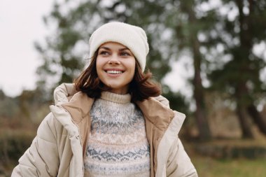 Açık hava portresi. Açık renk palto ve örgü şapkalı bir kadın. Doğal bir parkta serin bir günün tadını çıkarıyor. Moda, sağlık, seyahat ya da yaşam tarzı markalaşmasına uygun parlak, dostane bir ruh hali..