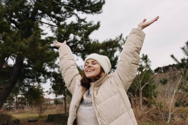 Açık havada mutlu bir kadın, kollarını kaldırıp kutlama yapıyor, kalın bir palto ve örgü şapka giyiyor, ağaçlarla çevrili ve açık manzaralı..