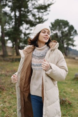 Havalı bir günde parkta dikilen kalın montlu, örgü ören, şapkalı bir kadınla sıradan bir kış moda sahnesi.