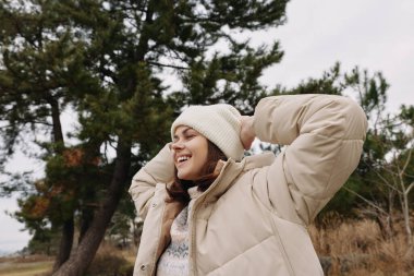 Sıcak bir palto ve bereyle serin havanın keyfini çıkaran bir kadının portresi, açık havada, gün ışığında ağaçların yanında, günlük moda ve sakin doğal manzara.