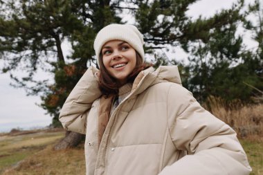 Açık havada bej bir ceket giyen ve doğal bir parkta sıcak bir bere takan, rahat kış kıyafetlerini ve rahat moda sahnelerini vurgulayan bir kadının portresi..
