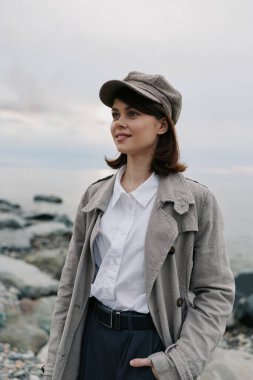 Tüvit ceketli ve şapkalı bir kadın, arkasında sakin bir deniz olan kayalık plajın yakınında poz veriyor. Doğal ışık, onun rahat ifadesini ve sonbahar stilini ön plana çıkarıyor, sıradan bir açık hava portresi konsepti..