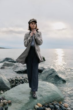 Paltolu ve şapkalı şık bir kadın bulutlu gökyüzü ve huzurlu deniz kenarında kayaların üzerinde poz veriyor. Sonbahar modası, doğa portresi, açık hava yaşam tarzı ve sakin sahil atmosferi kavramı..