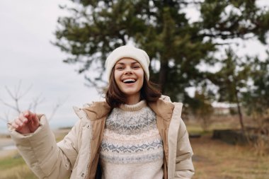 Serin havada dışarıda neşeli bir bayan, rahat bir ceket, ceket ve örgü şapkayla sarılmış, serin bir günde parkta yürürken parlak bir gülümsemeyi paylaşıyor..