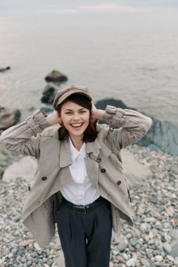 Samimi örülü hırka ve düz başlık giyen mutlu genç kadın sakin suyun yanındaki kayalık sahilde poz veriyor, neşeli bir ifade ve rahat bir duruş sergiliyor..