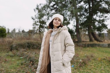 Açık bir alanda ağaçlarla, yumuşak bulutlu gökyüzü, dingin bir ruh hali, moda ve doğa birleşimi olan, yastıklı bir ceket, açık bir fular ve şapka giyen şık bir kadının açık hava portresi..