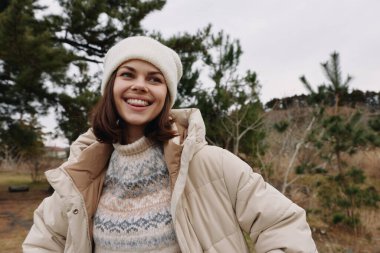 Soğuk hava kıyafetleri içinde neşeli bir kadının portresi, parkın içinde hiç bitmeyen yeşil ağaçlar, katmanlı giysiler ve sıcak aksesuarlar ile rahat bir açık hava yaşam tarzını yakalıyor. gevşemiş titreşim.