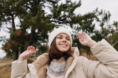 Açık havada bej ceketli ve kışlık ceketli gülümseyen bir kadın olarak sağlıklı, ışıl ışıl ve neşeli bir ifade. Günlük moda havası, doğal arka plan, samimi bir an ve açık hava yaşam tarzı.