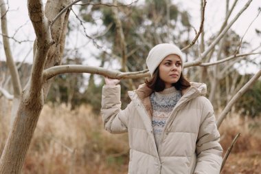 Sıcak ceketli genç bir kadın portresi ve kışlık bir parkta beyaz bir bere, etrafta çıplak dallar, yumuşak doğal ışık ve sakin açık hava atmosferi..