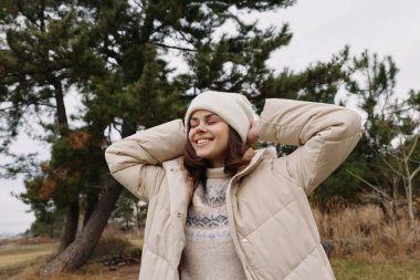 Sıcak bir palto ve örgü süveter giyen mutlu bir kadının sıradan dış portresi, rahat, cazibeli ve mevsimsel tarzda ağaçlarla doğal bir ortamda gevşek bir poz..