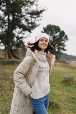 Yastıklı bir ceket ve açık havada rahat bir süveter giyen gülümseyen kadın, esinti ve yeşillikle dolu bir parkta poz veriyor, rahat bir stil yakalıyor, sıcaklığı ve samimi bir hayat tarzında doğal gün ışığını yakalıyor..