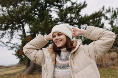 Açık hava sahnesinde paltolu gülümseyen bir kadın ve rüzgarlı bir tarlada örgü ören bir şapka, temiz havanın ve açık alanın tadını çıkarıyor.