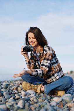 Doğanın tadını çıkaran sıradan bir fotoğrafçı, çakıl taşlı bir sahilde hazır kamera, çizmeli ve ekose gömlekli kadın neşeli bir ifadeyle poz veriyor, kıyı şeridi detaylarını ve dokularını belgesel tarzında belgeliyor..