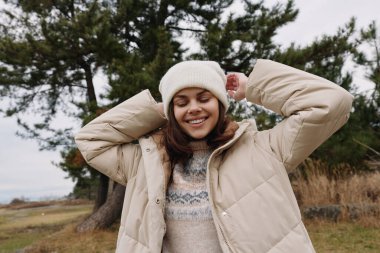 Sıcak bej ceket ve bere giyen neşeli bir kadının portresi. Açık alan ve ağaçlarla serin bir günün tadını çıkarıyor..