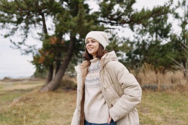 Kışlık giysiler içinde şık bir kadının açık hava portresi, bej ceket ve rahat bir kazak, yumuşak doğal ışık, kırsal alanda sonbahar ağaçlarının arasında rahat bir poz..