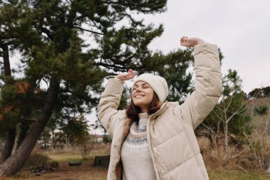 Dışarıdaki neşeli kadın kışlık bir ceket giyiyor ve örgü örüyor kutlama için kollarını kaldırıyor, çıplak ağaçların arasında gülümsüyor ve huzurlu bir park manzarası var..