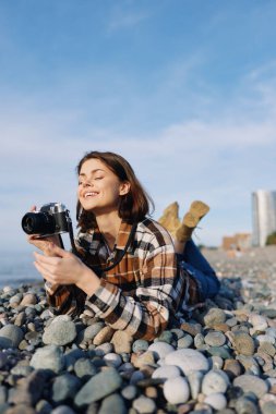Karaya vurmuş bir manzara, ekose gömlekli odaklanmış bir fotoğrafçı, elinde kamera, taşların arasında rahat bir poz, güneşli bir gün, yaratıcı keşif ve seyahat tarzı fotoğrafçılık.