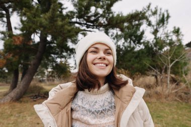 Paltolu ve bereli bir kadın portresi, parlak bir gülümseme, sakin bir doğa sahnesinde doğal ışık, günlük tarzını ve pozitif ruh halini ön plana çıkarıyor..
