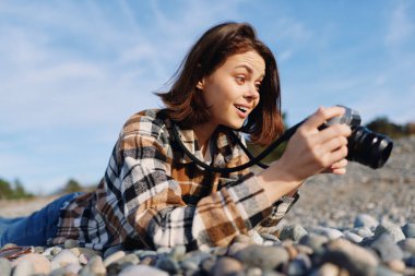 Sıradan bir kadın fotoğrafçı, kayalık bir sahilde canlı bir açık hava sahnesi yakalıyor, kamera hazır, güneş ışığı parlak, ekose gömlek giymiş, odaklanmış bakış, dinamik bir hobi anı.
