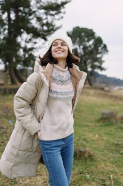 Açık hava portresi, kalın bir ceket giyen bir kadın, örgü süveter ve bere, açık alanda neşeli bir gülümseme, doğal ışık, rahat moda ve rahat açık hava havası..