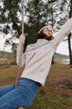 Salıncakta sallanan neşeli bir kadının açık hava portresi, rahat örgü bir kazak giyiyor, doğal bir ortamda, neşeli bir ruh hali ve tasasız bir duruşu var..