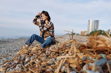 Sahil şeridinde sıradan bir kadın fotoğrafçı, deniz yosunlarını ve okyanus manzarasını odak ve merakla yakalıyor, açık hava macera havası ve güneşli gökyüzü altında seyahat ruhu..