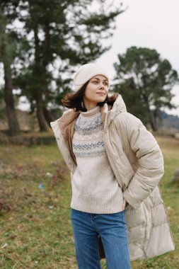 Açık havada, rahat bir kıyafetle, kabarık ceketle, örgü kazak ve şapkayla, rüzgarlı bir günde ağaçların ve çimenlerin arasında ellerle poz veren, kendinden emin ve doğal bir kadın..