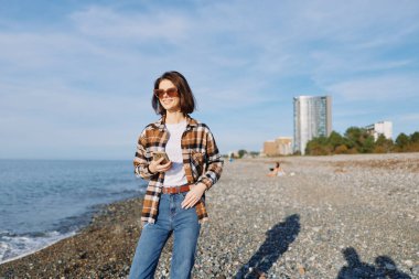 Sahilde rahat bir bayan gezgin, elinde bir telefon, ekose gömlek ve kot pantolon, güneş gözlüğü, dalgalı ve uzak şehir manzaralı aydınlık bir gün, sahil yürüyüşü sahnesi ve tasasız bir atmosfer..