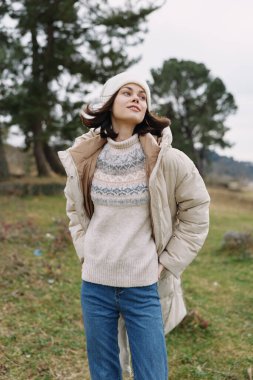 Sıcak bir süveter ve kabarık ceketli, kırsal bir parkta rahat bir poz ve esintili bir sonbahar atmosferi olan bir kadının açık hava portresi.