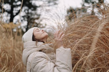 Açık hava rüzgarlı sahnesinde, uzun otların arasında sıcak bir palto giymiş bir kadın, dingin bir ifade, sonbahar ışığı, hafif bir esinti ve doğal dokular gizli doğa fotoğrafçılığını ve sakin bir ruh halini vurguluyor..