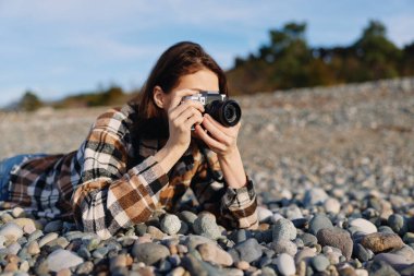 Kayalık bir sahilde, ekose elbiseli sıradan bir fotoğrafçı el kamerası, güneş ışığı sahnesi, desenli çakıl taşları ve otantik bir açık hava atmosferi ile kıyı manzarasını yakalar..