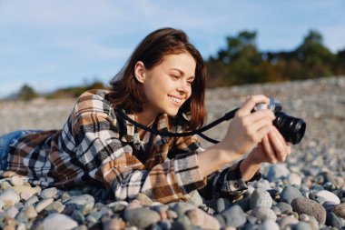 Kayalık bir plaj kamerasında kadın fotoğrafçı elinde. Kıyı şeridini fotoğraflıyor. Kaya üzerinde rahat bir poz.