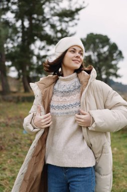 Açık hava portresi, kışın dış görünüşlü şık bir kadın, örgü süveter ve bere, ağaçlarla dolu çimenli bir alanda duruyor, rahat bir poz ve sakin bir ruh hali..